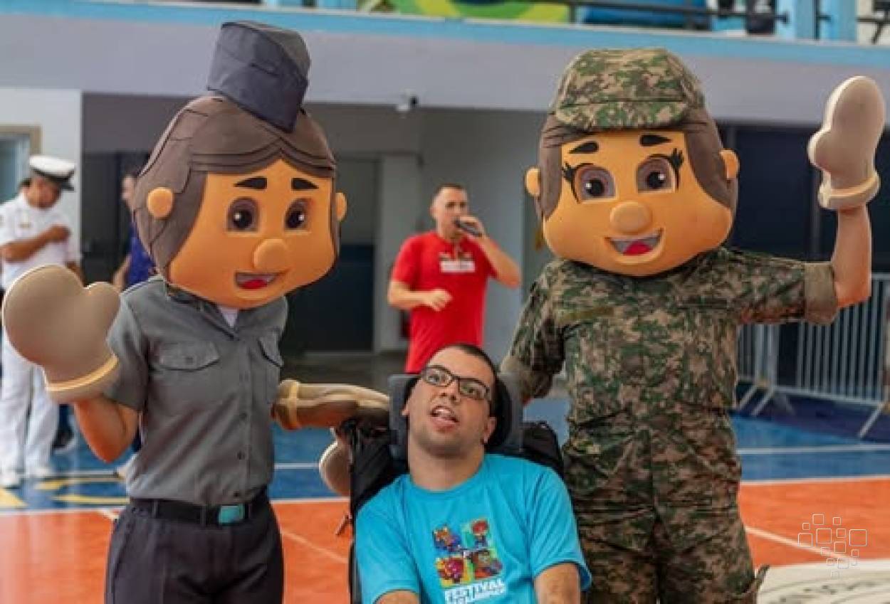 Participante em cadeira de rodas posa entre mascotes durante o Festival Paralímpico realizado no CEFAN